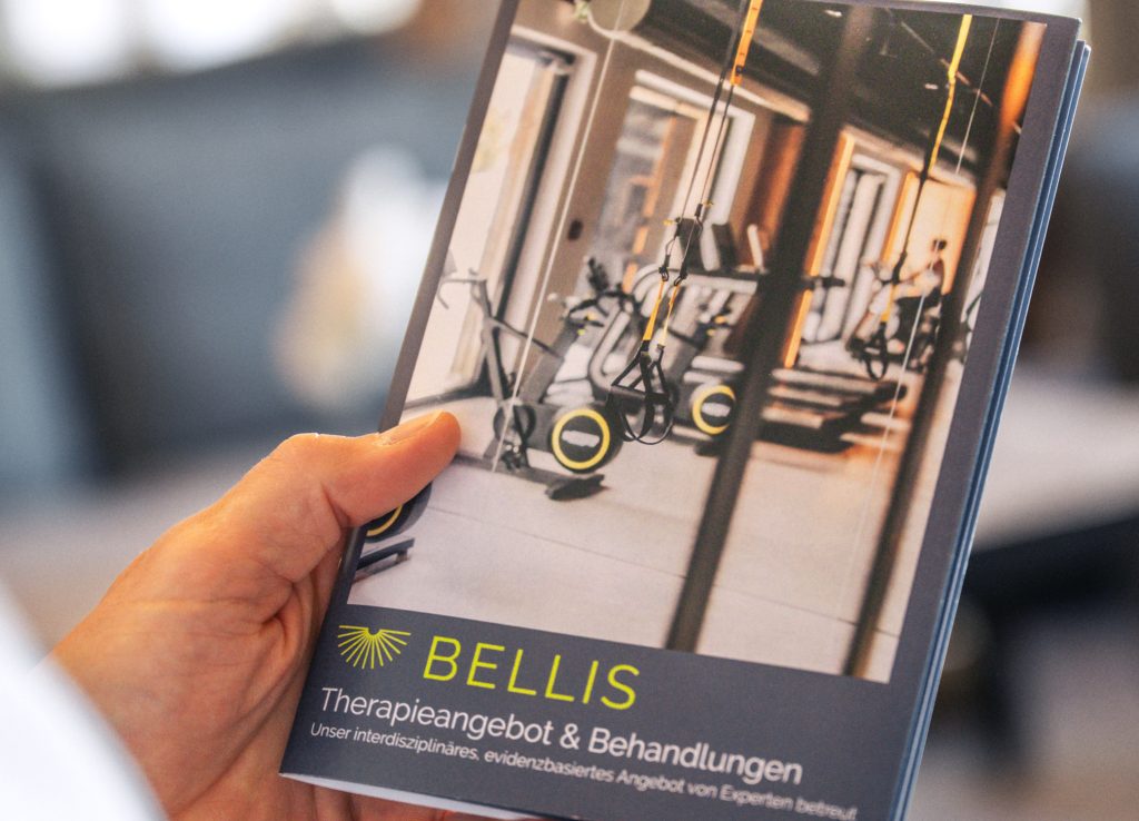 Broschüre Therapieangebot & Behandlungen mit Trainingsgeräten auf dem Titel, in der Hand gehalten, für BELLIS