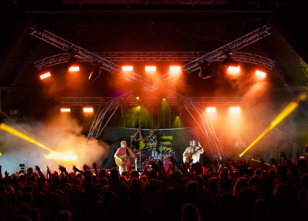 Band mit zwei Gitarristen und Schlagzeuger auf der Bühne in orangem Licht, davor Publikum mit erhobenen Händen