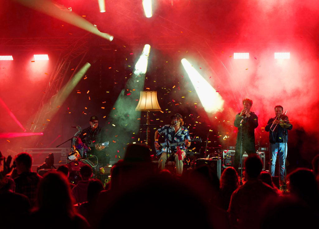 Band mit Trompete und Posaune auf der Bühne im roten Nebel, Konfetti und Stehlampe, davor das Publikum, BK Open Air 2025
