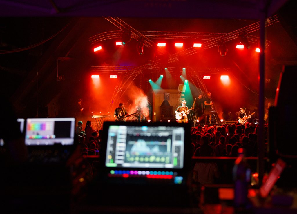 Blick über Bildschirme der Bühnentechnik auf Band und Publikum im roten Licht, BK Open Air 2025