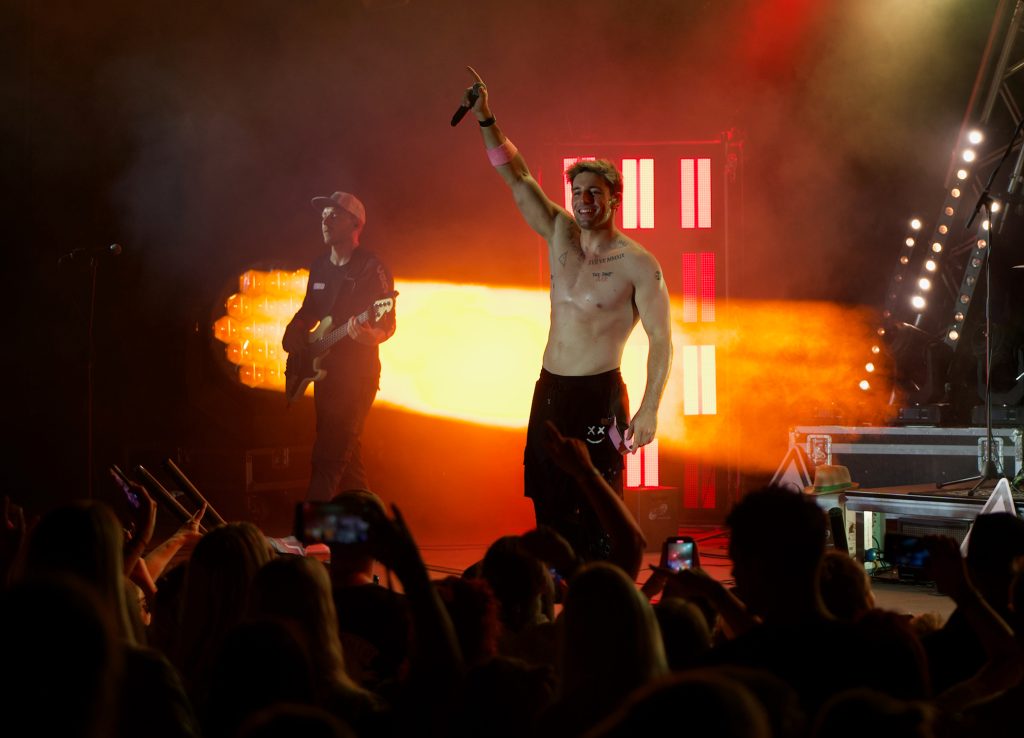 Sänger Alexander Eder mit erhobenem Arm und Mikrofon auf der Bühne, links ein Bassist, oranges Licht, BK Open Air 2025