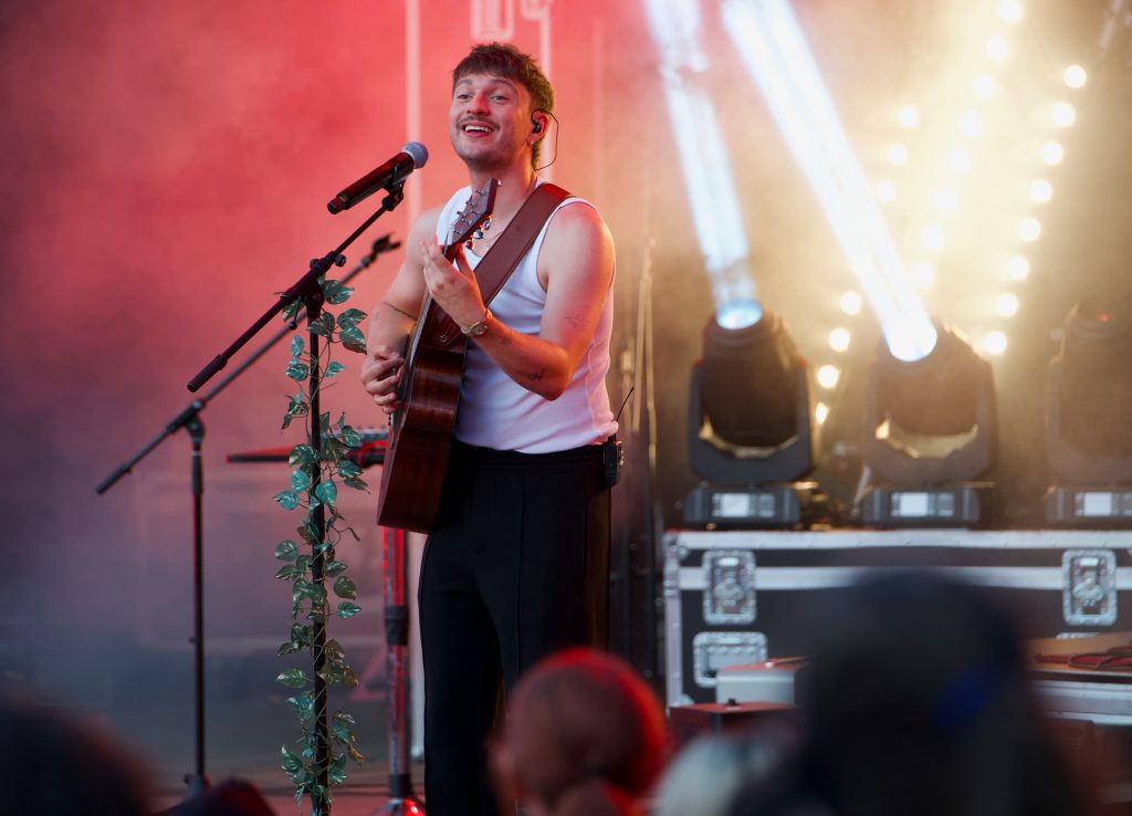 Lächelnder Sänger Rian mit Gitarre am Mikrofonständer mit Blätterranke, helle Scheinwerfer im Hintergrund, BK Open Air 2025