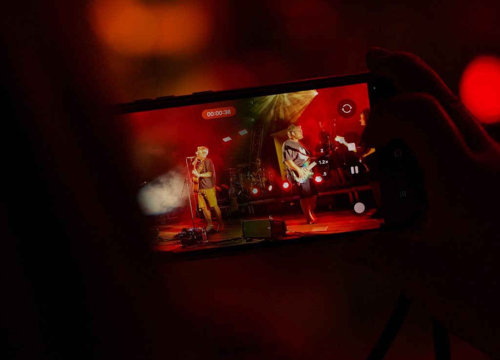 Hand hält ein Smartphone und filmt Musiker auf der roten Bühne beim BK Open Air 2025