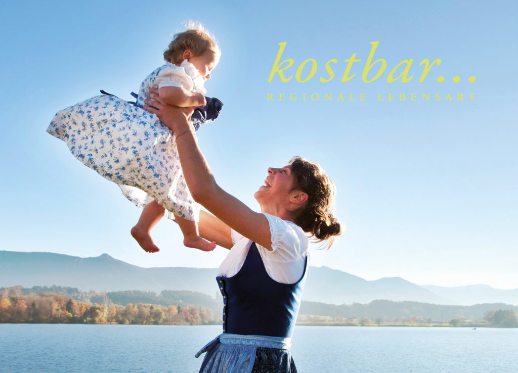 Frau in Tracht hebt ein Kind vor See und Bergen hoch, Schriftzug kostbar Regionale Lebensart, für kostbar