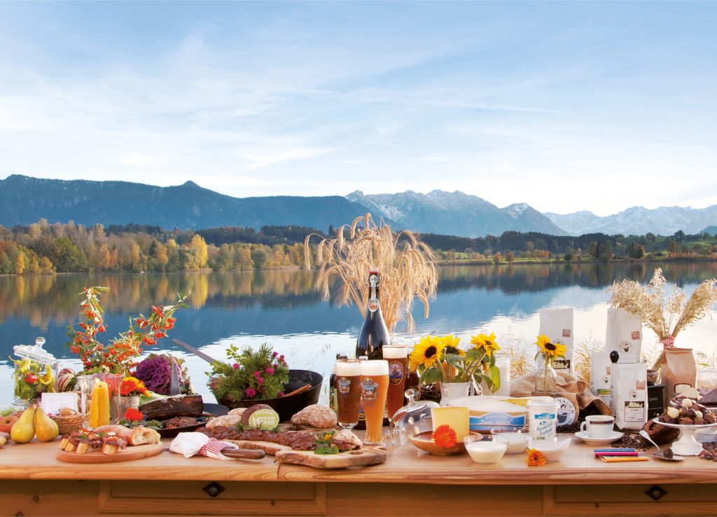 Gedeckter Tisch am See mit Bier, Fleisch, Gemüse, Joghurt, Kaffee und Pralinen vor Bergen für kostbar