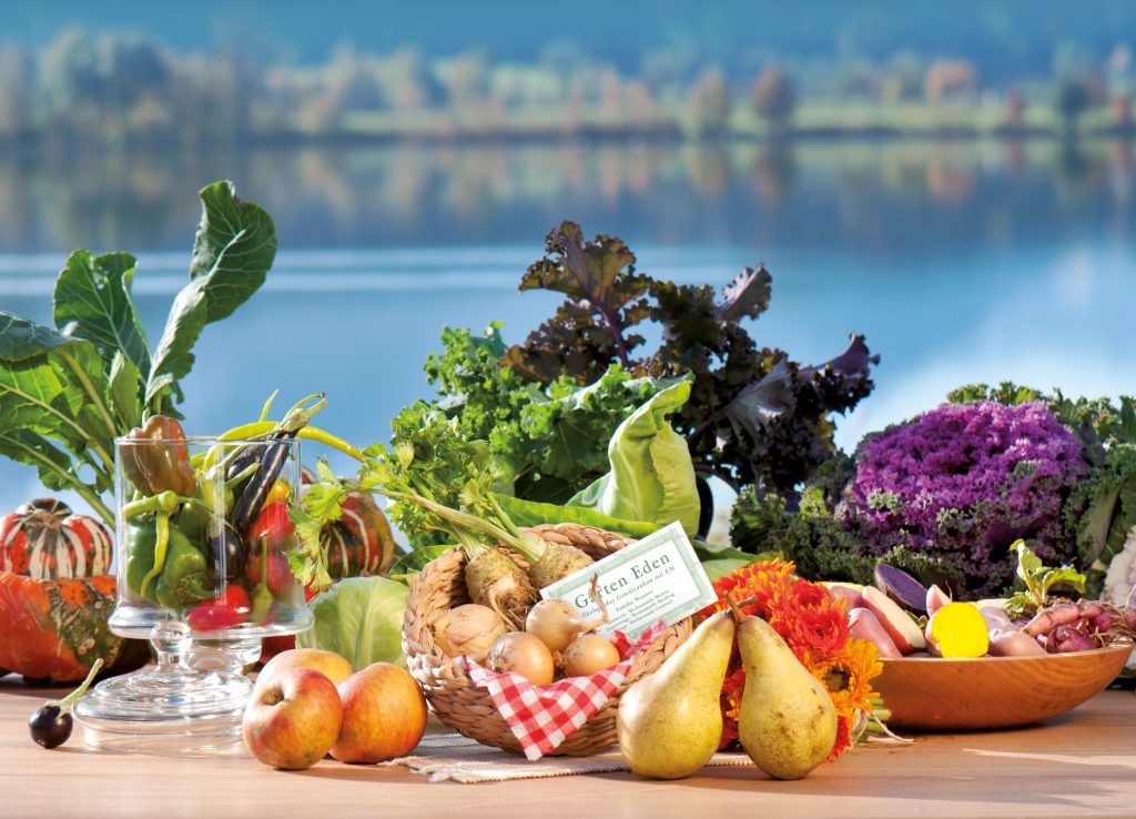 Gemüse, Äpfel, Birnen und Blumen auf Holztisch vor See, im Korb eine Karte Garten Eden, für kostbar