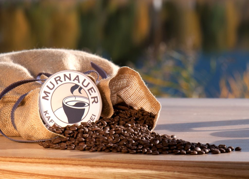 Umgekippter Jutesack mit Kaffeebohnen und Logo Murnauer Kaffeerösterei auf Holztisch vor See für kostbar