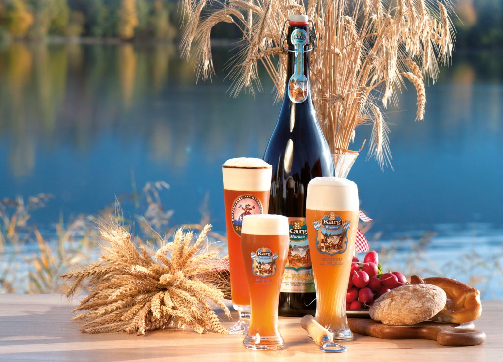 Drei Gläser und eine Flasche Karg Weißbier mit Weizenähren, Radieschen, Brot und Brezel vor See für kostbar