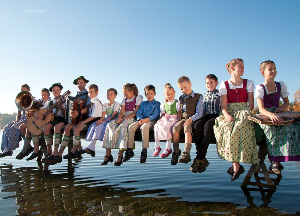 Dreizehn Kinder und Jugendliche in Tracht mit Tuba, Gitarre, Harmonika und Zither sitzen am Steg für kostbar